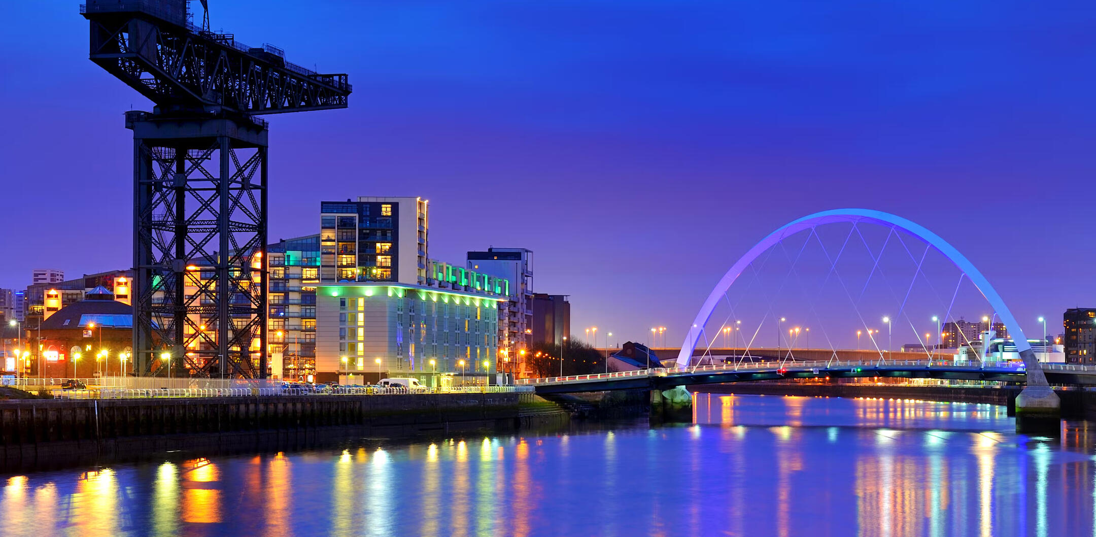 A night photo of the River Clyde. There is the Finniston crane on the left and the squinty bridge on the right. There are lots of street lights and coloured lights reflecting off the river.